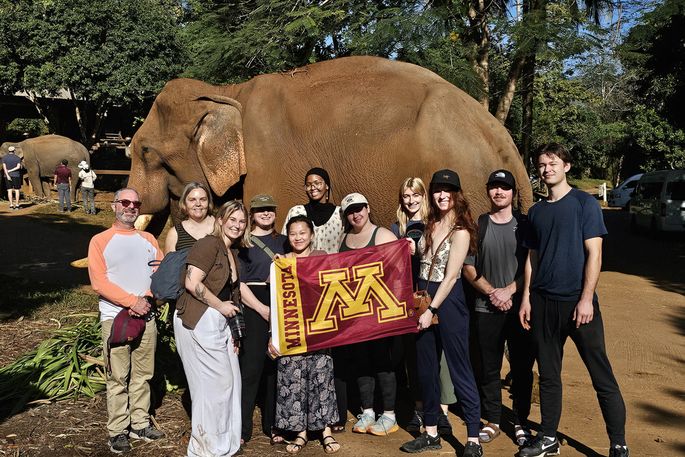 A group of students hold a Block M flag and pose for a photo in front of an elephant in Thailand