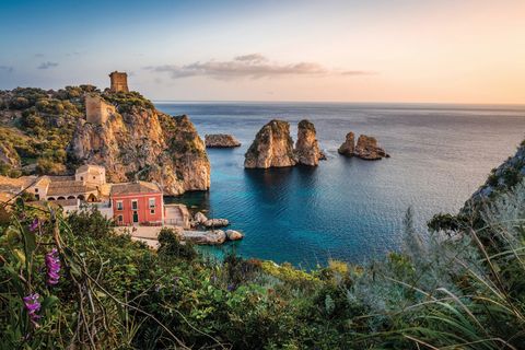 The sun sets over an Italian house and the blue-green seaside. Giant rocks emerge from the water beyond.