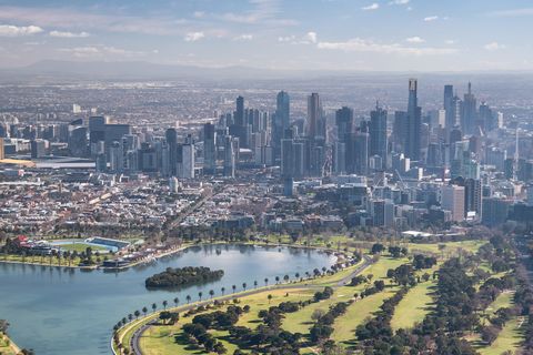 The sun shines brightly on the city of Melbourne, with skyscrapers towering over smaller buildings and a lush green area on the waterfront 