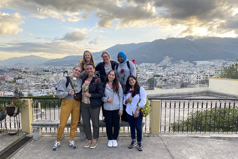 A group of students hold roses while posing for a photo in front of a city and mountains in Ecuador