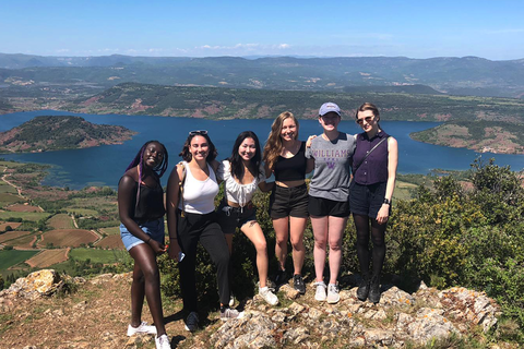 A group of students poses for a picture on top of a lush and rugged hill, with a glimmering blue lake in the background